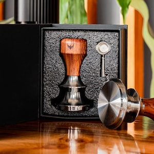 May include: A black box with a wooden coffee tamper, a small metal tamper, and a metal scoop inside. The box is open and the items are visible.