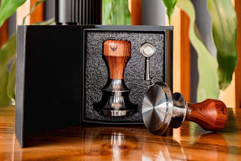 May include: A black box with a wooden coffee tamper, a small metal tamper, and a metal scoop inside. The box is open and the items are visible.