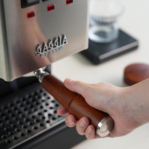 May include: A stainless steel Gaggia Classic espresso machine with a wooden handle portafilter. The machine has three illuminated buttons and the Gaggia logo. The portafilter is held by a person's hand.