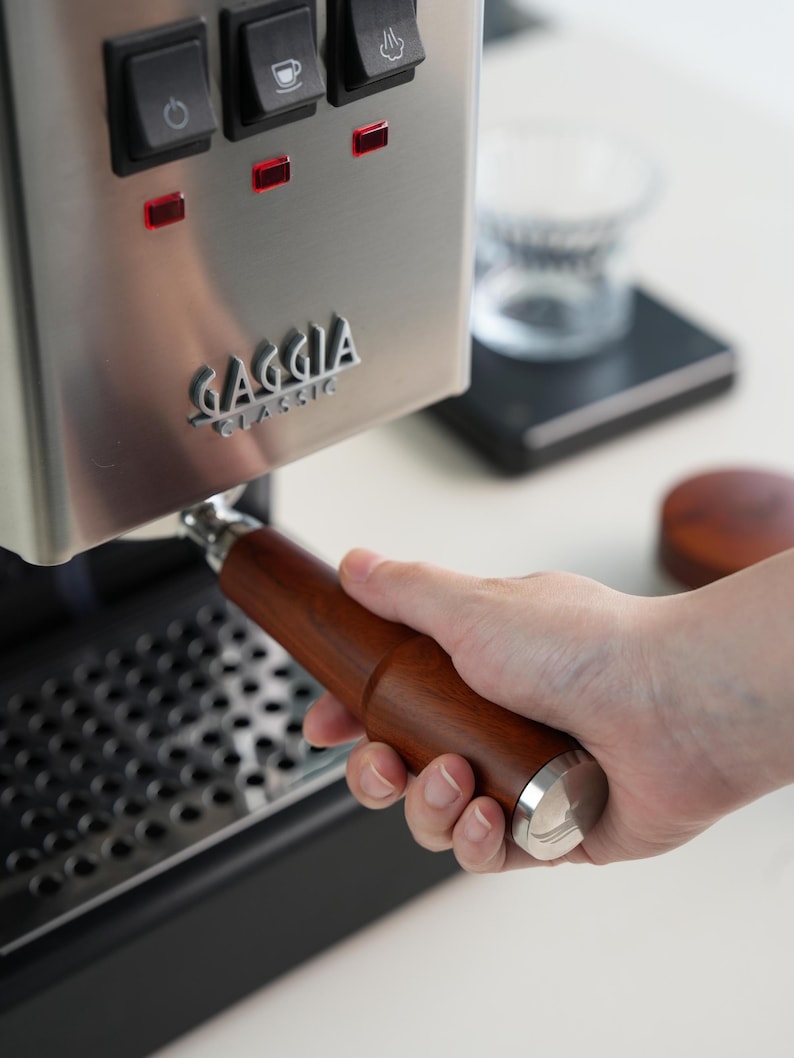 May include: A stainless steel Gaggia Classic espresso machine with a wooden handle portafilter. The machine has three illuminated buttons and the Gaggia logo. The portafilter is held by a person's hand.