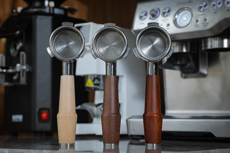 May include: Three espresso portafilters with wooden handles. The handles are made of different types of wood: light wood, dark wood, and reddish wood.