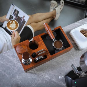 Puede incluir: Una estación de espresso de madera con un portafiltro, un tamper y otros accesorios. Una balanza blanca con granos de café está cerca. Una persona sostiene una taza de café con arte latte.