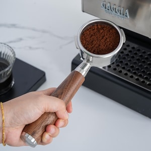 May include: A stainless steel portafilter with a wooden handle, filled with ground coffee. The portafilter is held near a Gaggia espresso machine. The handle is a dark brown colour.