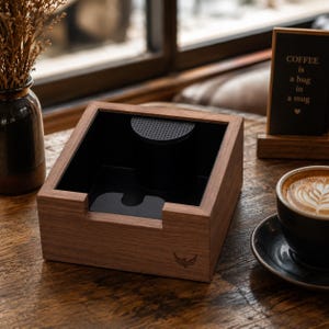 Puede incluir: Una estación de café de madera con interior negro y filtro metálico. La estación tiene un recorte para un portafiltro y un pequeño logotipo. Un letrero dice "El café es un abrazo en una taza". También hay una taza de café con arte latte.