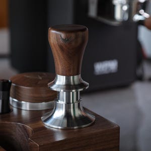 May include: A wooden and stainless steel espresso tamper with a walnut handle. The tamper is sitting on a wooden surface.