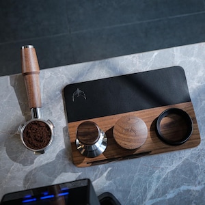 May include: A wooden coffee tamper station with a black leather mat, three wooden tamper bases, and a metal tamper. The station is on a gray marble surface.