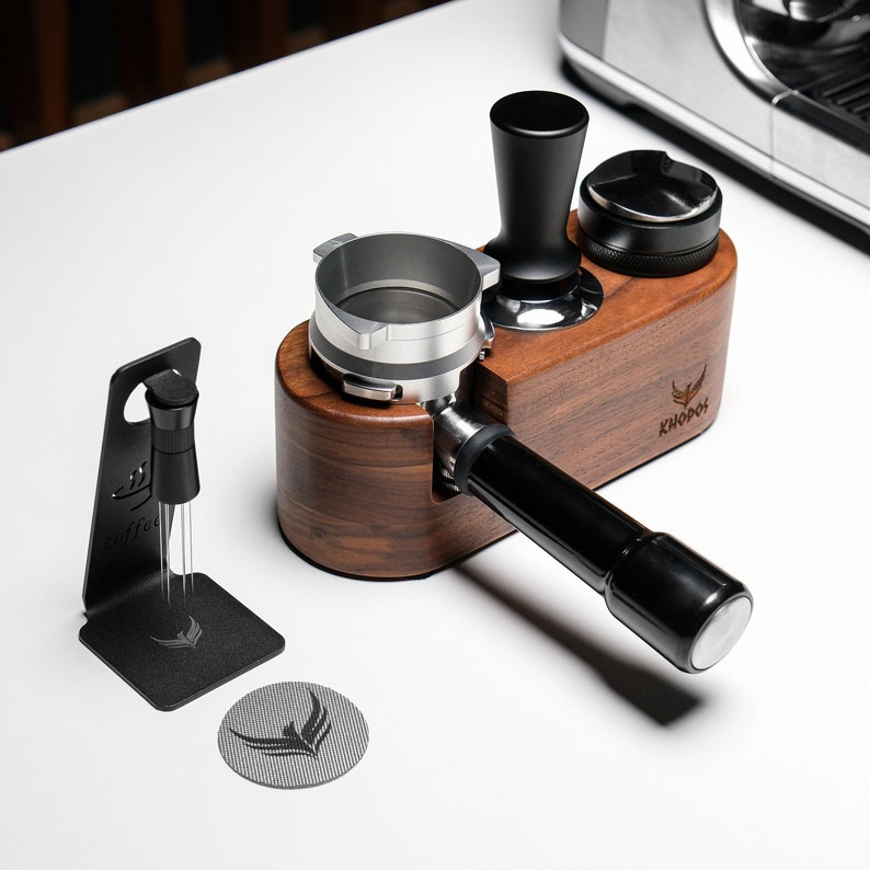 May include: A wooden espresso stand with a black tamper, a portafilter, and a black and silver tool. The stand has the word "KHODOS" printed on it.