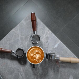 May include: A close-up of a latte art design in a white ceramic cup, surrounded by three espresso tampers with wooden handles. The tampers are arranged in a triangular shape around the cup.