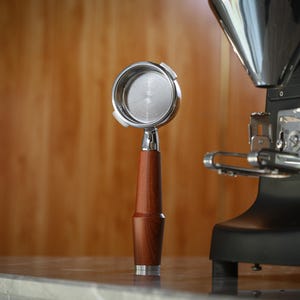 May include: A stainless steel portafilter with a wooden handle is positioned in front of a black coffee machine. The portafilter has a polished metal filter basket. The wooden handle is a rich, dark brown colour.
