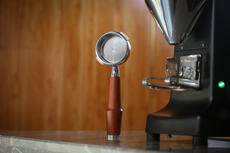 May include: A stainless steel portafilter with a wooden handle is positioned in front of a black coffee machine. The portafilter has a polished metal filter basket. The wooden handle is a rich, dark brown colour.