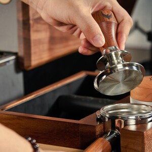 May include: A wooden box with a built-in tamper for coffee grounds. The tamper has a wooden handle and a stainless steel base. The box is open and the tamper is being placed inside.