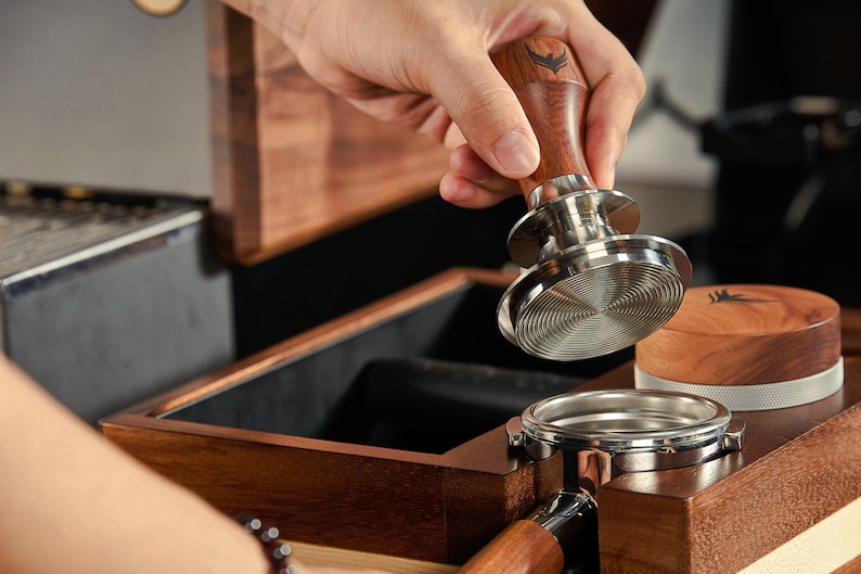 May include: A wooden box with a built-in tamper for coffee grounds. The tamper has a wooden handle and a stainless steel base. The box is open and the tamper is being placed inside.