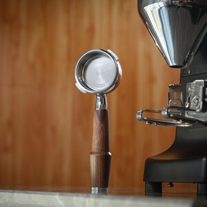 May include: A coffee portafilter with a wooden handle and a stainless steel filter, next to a black coffee grinder. The portafilter is made of stainless steel and wood. The grinder is black with a silver hopper.
