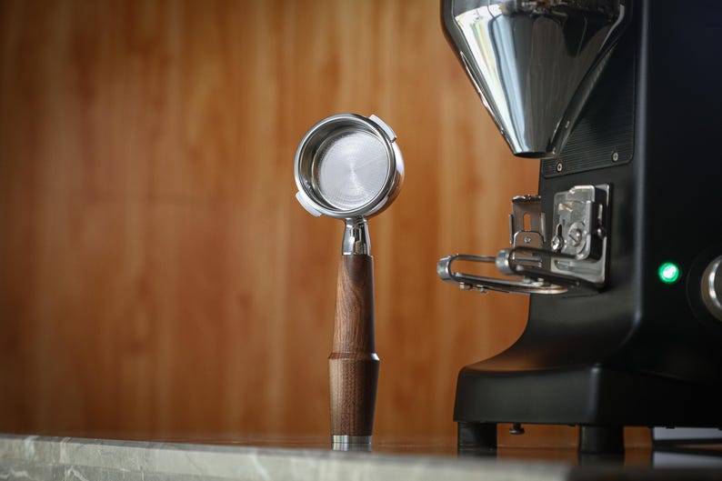 May include: A coffee portafilter with a wooden handle and a stainless steel filter, next to a black coffee grinder. The portafilter is made of stainless steel and wood. The grinder is black with a silver hopper.