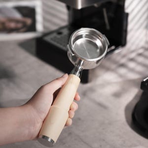May include: A stainless steel portafilter with a light wood handle. The portafilter is held in front of a coffee machine. The handle is cylindrical and has a silver metal base. The portafilter is empty and ready for coffee grounds.