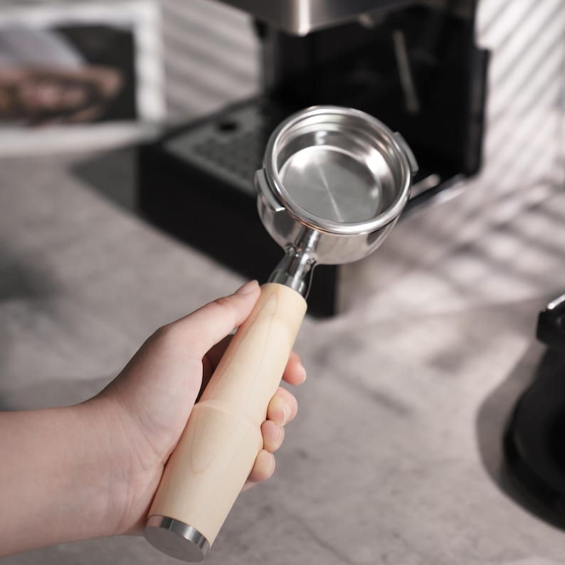 May include: A stainless steel portafilter with a light wood handle. The portafilter is held in front of a coffee machine. The handle is cylindrical and has a silver metal base. The portafilter is empty and ready for coffee grounds.