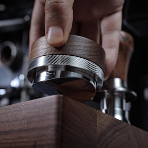 Peut inclure: Gros plan sur un tamper à café en bois et en métal avec un anneau argenté et le chiffre "4" sur le côté. Le tamper est tenu par une main et repose sur une surface en bois.