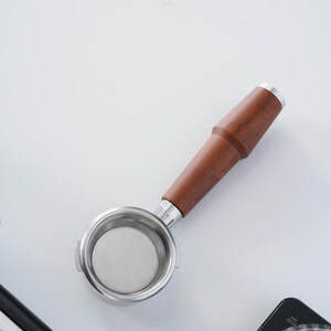 May include: A stainless steel portafilter with a wooden handle. The portafilter has a circular basket with a fine mesh filter. The handle is a dark brown colour with a silver accent at the end.