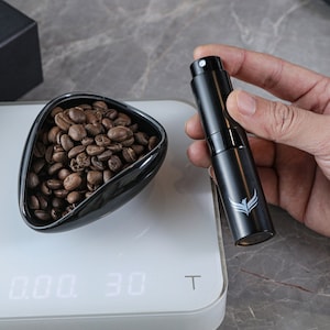 May include: A black digital scale with a white display showing "0.00 30 T". A black spray bottle with a white logo is held in a hand above the scale. A bowl of coffee beans is on the scale.