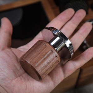 Puede incluir: Un tamper de café con mango de madera y base de acero inoxidable. El mango es de color marrón oscuro y tiene ranuras verticales. La base es plateada y tiene grabado el texto "DOS Echo Tamper". El tamper está diseñado para prensar el café molido.