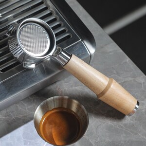 May include: A stainless steel portafilter with a wooden handle, used for making espresso. The portafilter is sitting on top of a silver espresso machine.