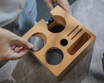 May include: A wooden coffee pod holder with four compartments for coffee pods. The holder has a small compartment for a coffee scoop and a larger compartment for a coffee bag. The holder is made of light wood and has a natural finish.