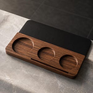 Puede incluir: Una bandeja rectangular de madera con tres huecos circulares y una sección superior negra. La bandeja está hecha de madera oscura y tiene un acabado liso. La bandeja está diseñada para guardar objetos pequeños.