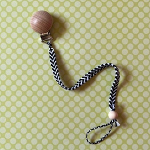 May include: A black and white chevron patterned baby pacifier clip with a wooden bead and a wooden teether. The clip is attached to a light green polka dot background.