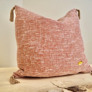 May include: A soft, peach-colored throw pillow with a subtle woven texture and tassel accents. The pillow has a small white label with text.