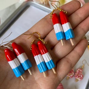 Puede incluir: Pendientes artesanales con dise&ntilde;o de helado de paleta en rojo, blanco y azul. Cada pendiente presenta un dise&ntilde;o de mini helado de paleta con un palito de madera y ganchos dorados. Un divertido accesorio veraniego.