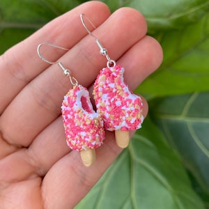 Puede incluir: Un par de pendientes de paleta. Cada pendiente tiene la forma de una paleta rosa con chispas blancas y amarillas. Las paletas est&aacute;n unidas a ganchos para la oreja de color plateado.