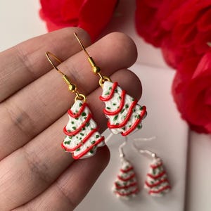 May include: Christmas tree earrings with gold-coloured hooks. The earrings are white with red and green accents, resembling a decorated Christmas tree. Two smaller pairs of earrings are also visible.