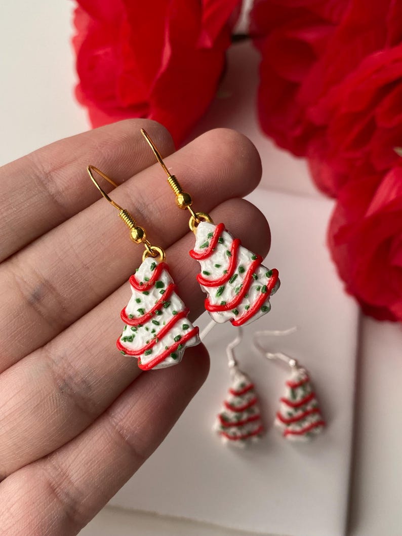 May include: Christmas tree earrings with gold-coloured hooks. The earrings are white with red and green accents, resembling a decorated Christmas tree. Two smaller pairs of earrings are also visible.