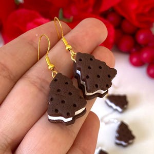 May include: Novelty earrings in the shape of chocolate cookie sandwiches. The earrings have a dark brown cookie exterior with white cream filling and gold-coloured ear wires. Additional earrings are visible in the background.