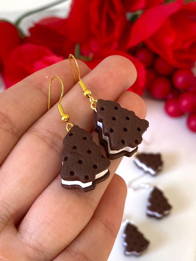 May include: Novelty earrings in the shape of chocolate cookie sandwiches. The earrings have a dark brown cookie exterior with white cream filling and gold-coloured ear wires. Additional earrings are visible in the background.