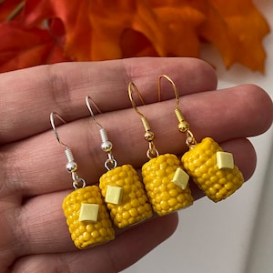 May include: Four pairs of corn on the cob earrings. Two pairs have gold hooks and two pairs have silver hooks. Each corn on the cob is yellow with a small square of white butter on top.