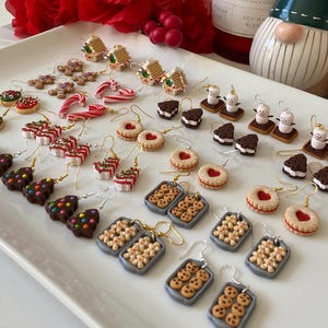 May include: A selection of festive earrings featuring miniature gingerbread houses, gingerbread men, candy canes, cookies, and Christmas trees. The earrings are in shades of red, white, brown, and green. The earrings are presented on a white tray, perfect for the holiday season.