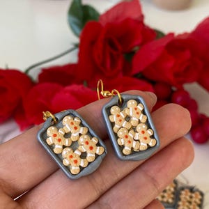 May include: Handmade gingerbread man earrings. Each earring depicts a miniature grey baking tray filled with gingerbread men. The gingerbread men are tan with white icing and red accents. The earrings have gold-coloured hooks.
