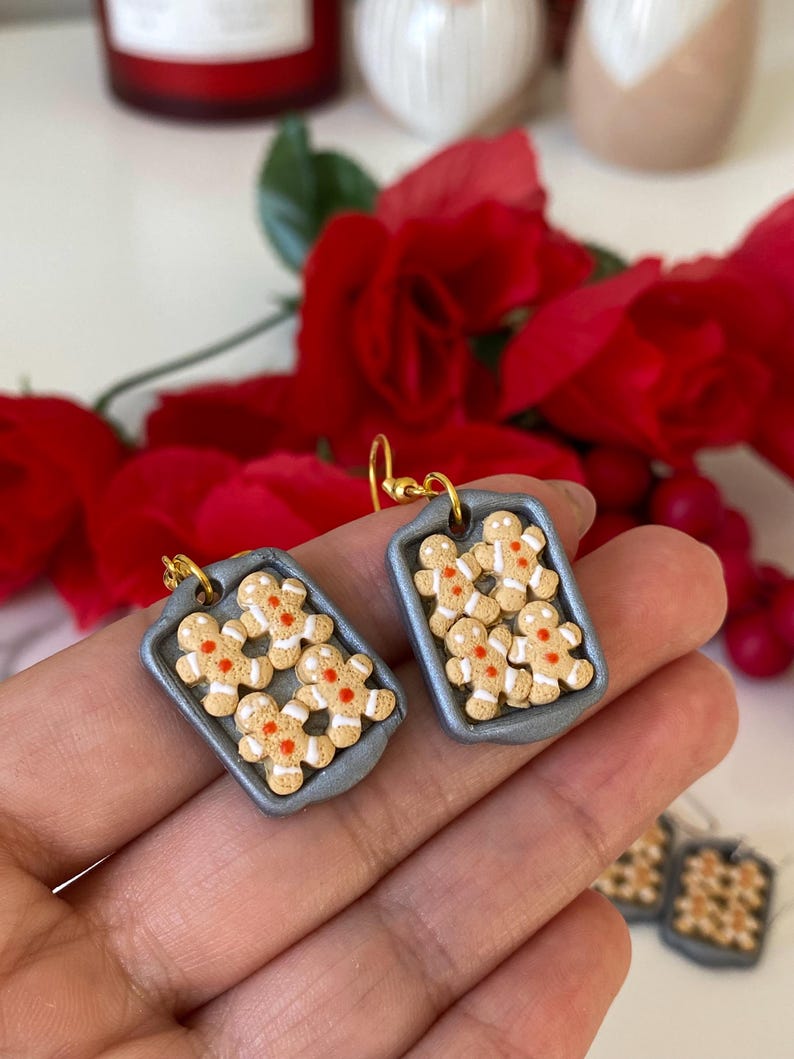 May include: Handmade gingerbread man earrings. Each earring depicts a miniature grey baking tray filled with gingerbread men. The gingerbread men are tan with white icing and red accents. The earrings have gold-coloured hooks.