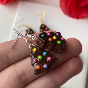 May include: A pair of handmade chocolate-coloured Christmas tree earrings with colourful dot decorations. The earrings have silver-coloured hooks. The background includes a red rose and a white surface.