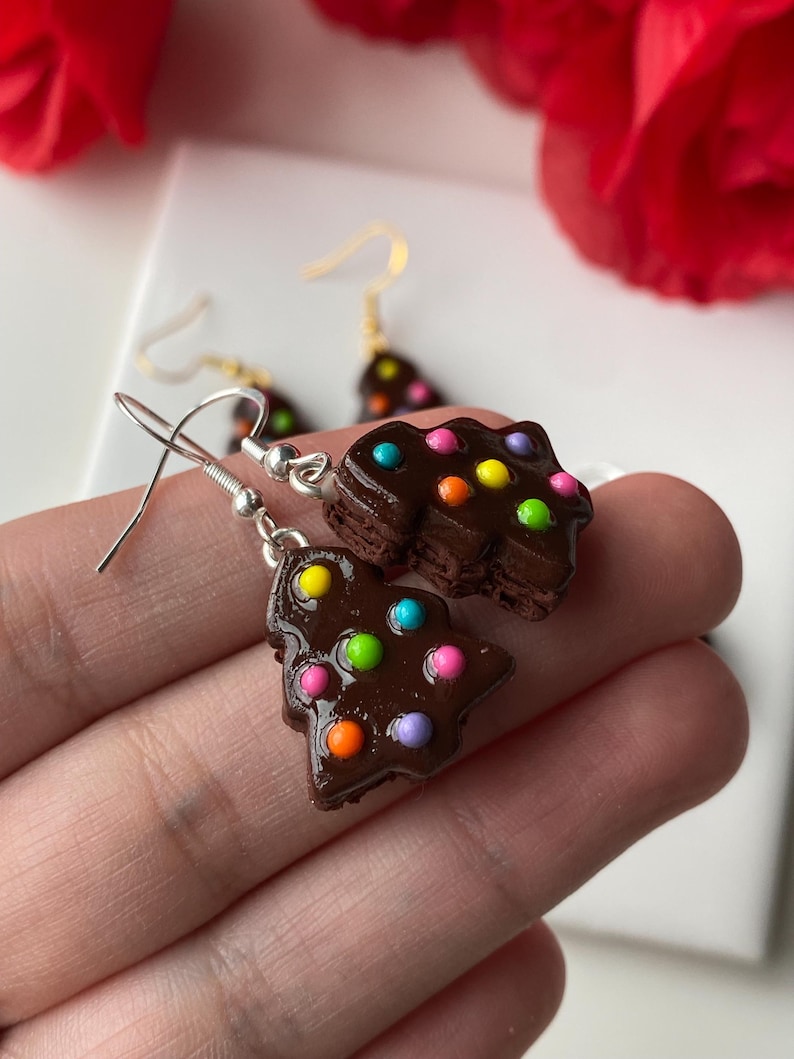 May include: A pair of handmade chocolate-coloured Christmas tree earrings with colourful dot decorations. The earrings have silver-coloured hooks. The background includes a red rose and a white surface.