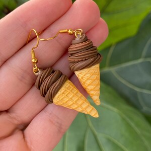 Puede incluir: Pendientes hechos a mano con forma de conos de helado en miniatura. Cada pendiente tiene una base de gofre rematada con helado en espiral de color chocolate. Los pendientes cuelgan de ganchos dorados.