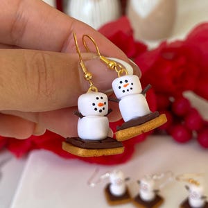 May include: A pair of novelty snowman earrings. Each earring depicts a white marshmallow snowman with black dot eyes and an orange nose, resting on a biscuit base. The earrings have gold-coloured hooks and are a fun, festive accessory.