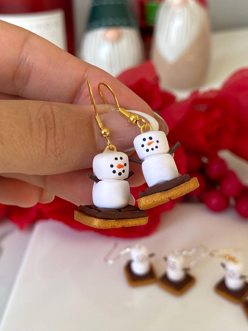 May include: A pair of novelty snowman earrings. Each earring depicts a white marshmallow snowman with black dot eyes and an orange nose, resting on a biscuit base. The earrings have gold-coloured hooks and are a fun, festive accessory.