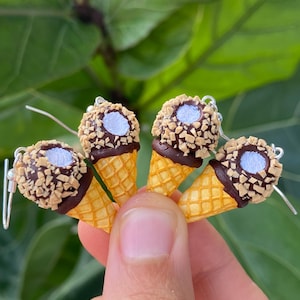 Puede incluir: Pendientes de cono de helado en miniatura. Cada pendiente presenta un cono de gofre cubierto con chocolate, nueces y una bola de helado de vainilla. Los pendientes est&aacute;n sostenidos por los dedos de una persona contra un fondo verde y frondoso.