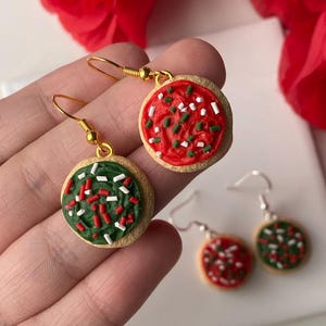 May include: Cookie-shaped earrings with gold-coloured hooks. One earring has red icing with white and green sprinkles, the other has green icing with red and white sprinkles. A festive accessory.