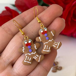 May include: Gingerbread man earrings. The earrings are brown with white icing details, red mouths, purple buttons, and blue eyes. They are suspended from gold-coloured hooks. A festive accessory.