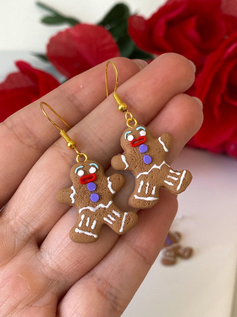 May include: Gingerbread man earrings. The earrings are brown with white icing details, red mouths, purple buttons, and blue eyes. They are suspended from gold-coloured hooks. A festive accessory.