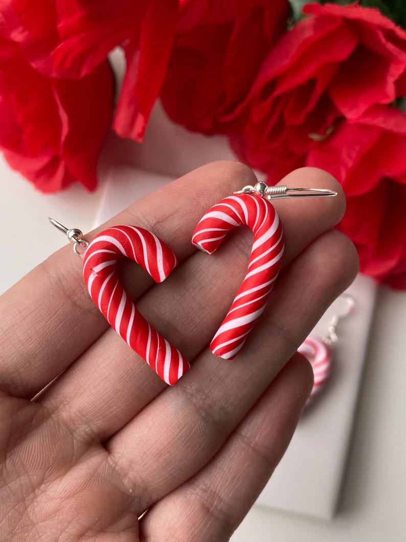 May include: A pair of candy cane earrings. Each earring is a red and white striped candy cane shape with a silver hook. The earrings are displayed against a background of red flowers.