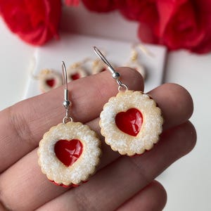 May include: A pair of dangle earrings resembling heart-shaped jammy dodgers. The earrings feature a beige biscuit base, a red heart centre, and a dusting of white powder. They hang from silver hooks.